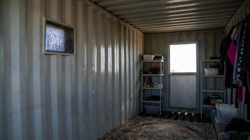 Interior of a shipping container home showing the narrow 7.5-foot width constraint with rug, shelving, and clothing rack